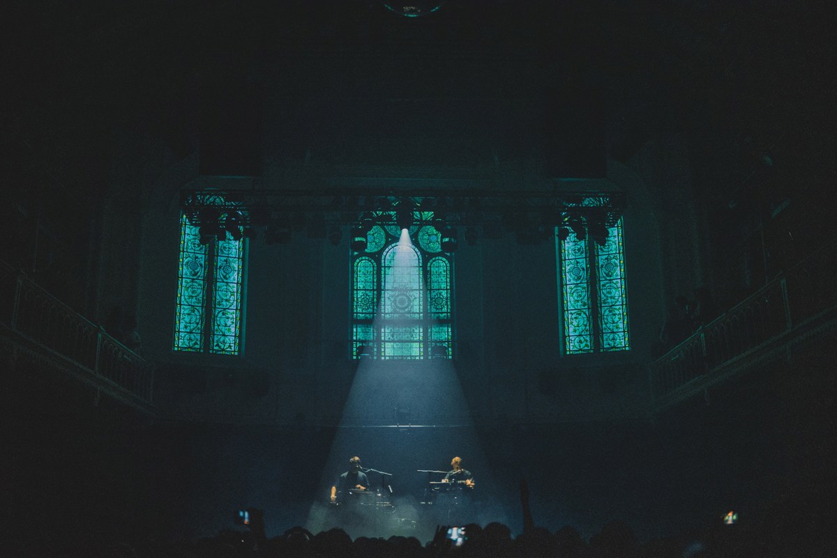Een foto van achterin de zaal van Paradiso. Een spot richt van bovenin de zaal op het podium. We zien de kerkramen van Paradiso.