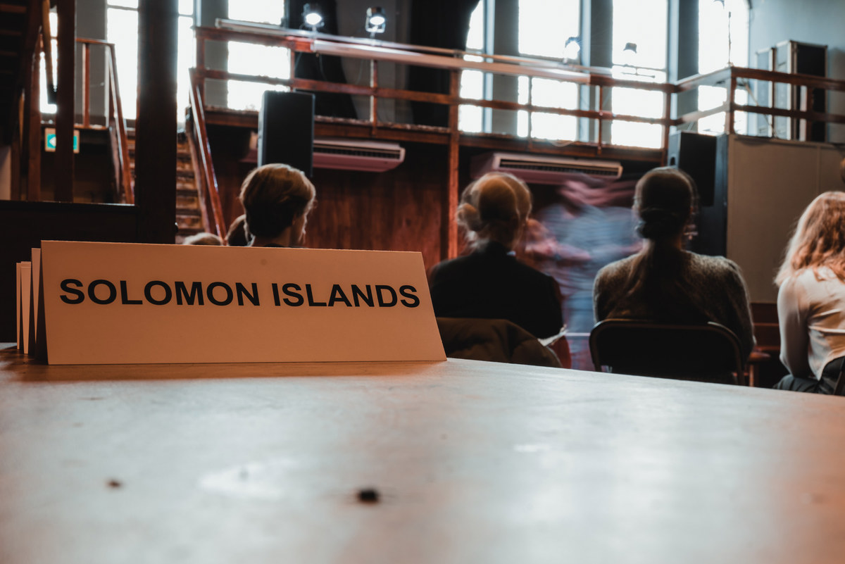 Uitleg op Flip the Script. Een man in beweging achter een bordje met 'SOLOMON ISLANDS'