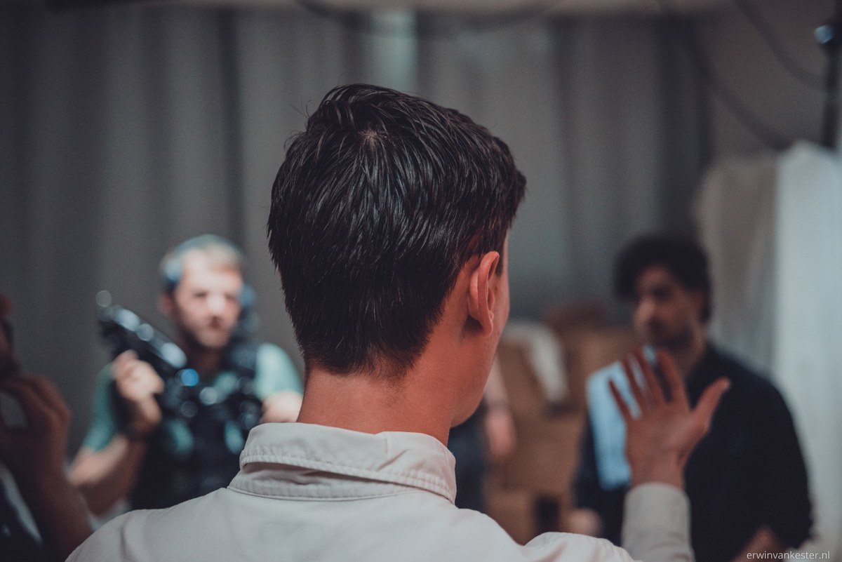 Achterhoofd van een man op de voorgrond terwijl een cameraman met schoudercamera en een gesticulerende man op de achtergrond scherp in beeld verschijnen.