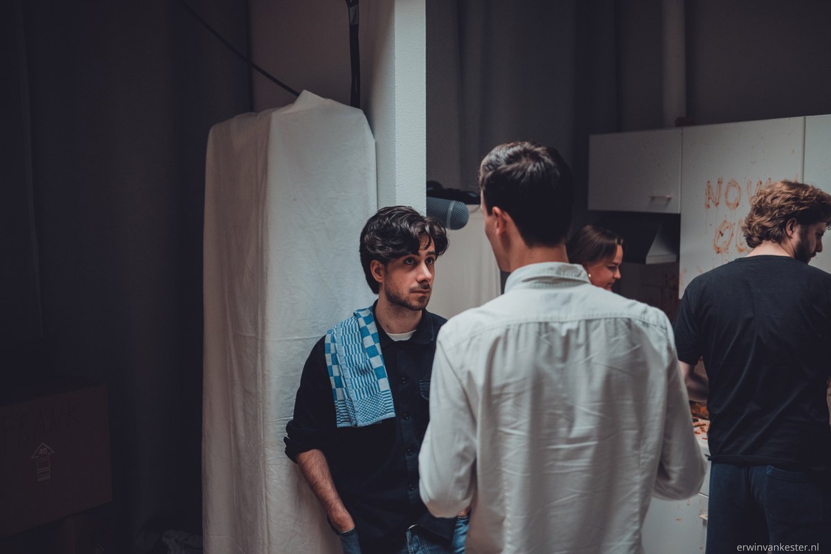 Twee jonge mannen staan in gesprek in een sfeervol verlichte studioruimte, terwijl op de achtergrond enkele andere personen bij een tafel bezig zijn.