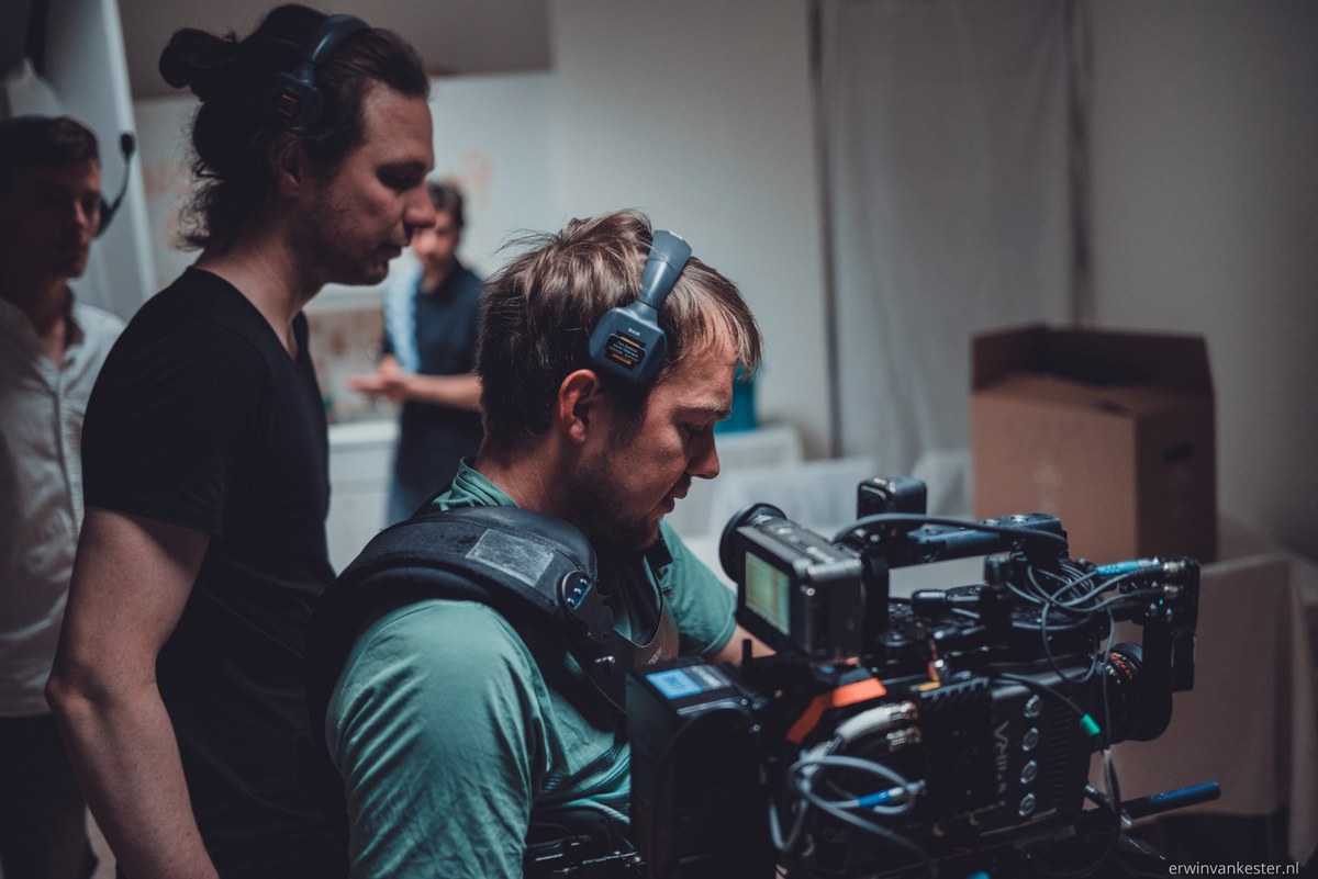Twee filmcrewleden met koptelefoons op werken samen achter een professionele filmcamera tijdens een opname in een sober ingerichte ruimte.