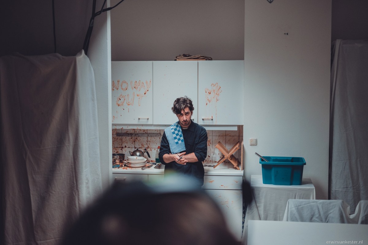 Man met blauwe theedoek over de schouder staat in een keuken met vaat, terwijl op de kastjes in rode letters No Way Out is geschreven.