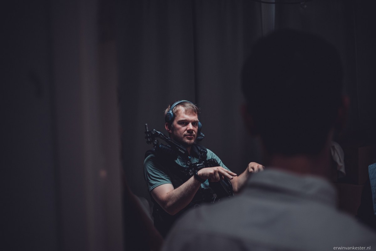Man met koptelefoon in een groen shirt zit gebarend te praten met een persoon op de voorgrond in een sfeervol verlichte studio.