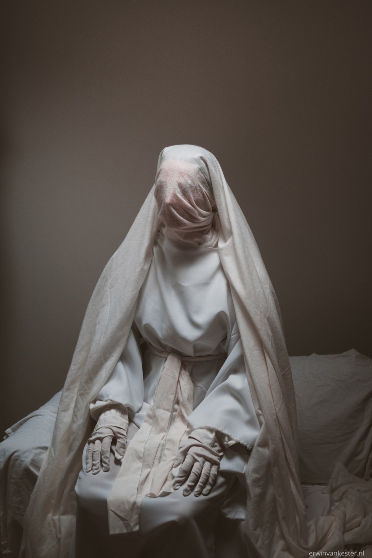 Een in witte doeken en een sluier gehulde figuur zit met de handen op de knieën op een bed, tegen een sobere bruine muur.
