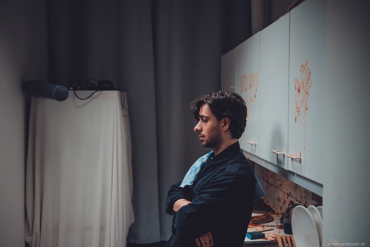 Man met gekruiste armen staat in gedachten verzonken naast keukenkastjes met rode vlekken, in een schemerige ruimte met grijze gordijnen.