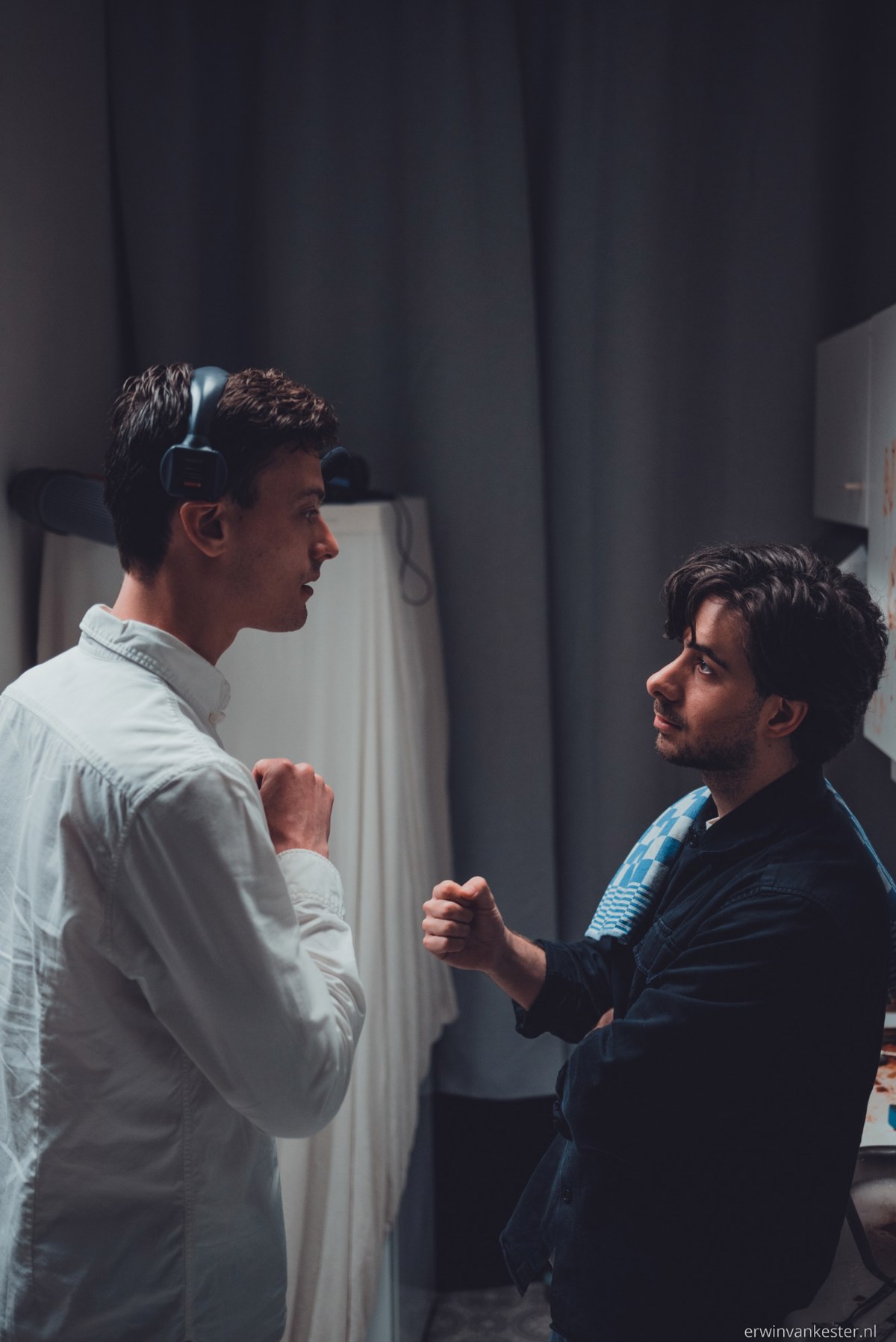 Twee mannen staan tegenover elkaar in gesprek, waarbij de linker een headset draagt en de rechter zijn hand gebald voor zich houdt in een donkere ruimte.