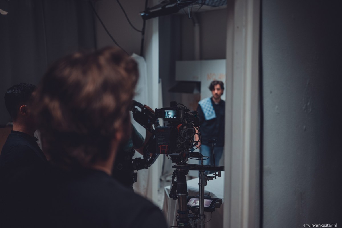 Op de voorgrond bedient een crewlid een filmcamera terwijl op de achtergrond een acteur met baard scherpgesteld in beeld staat.