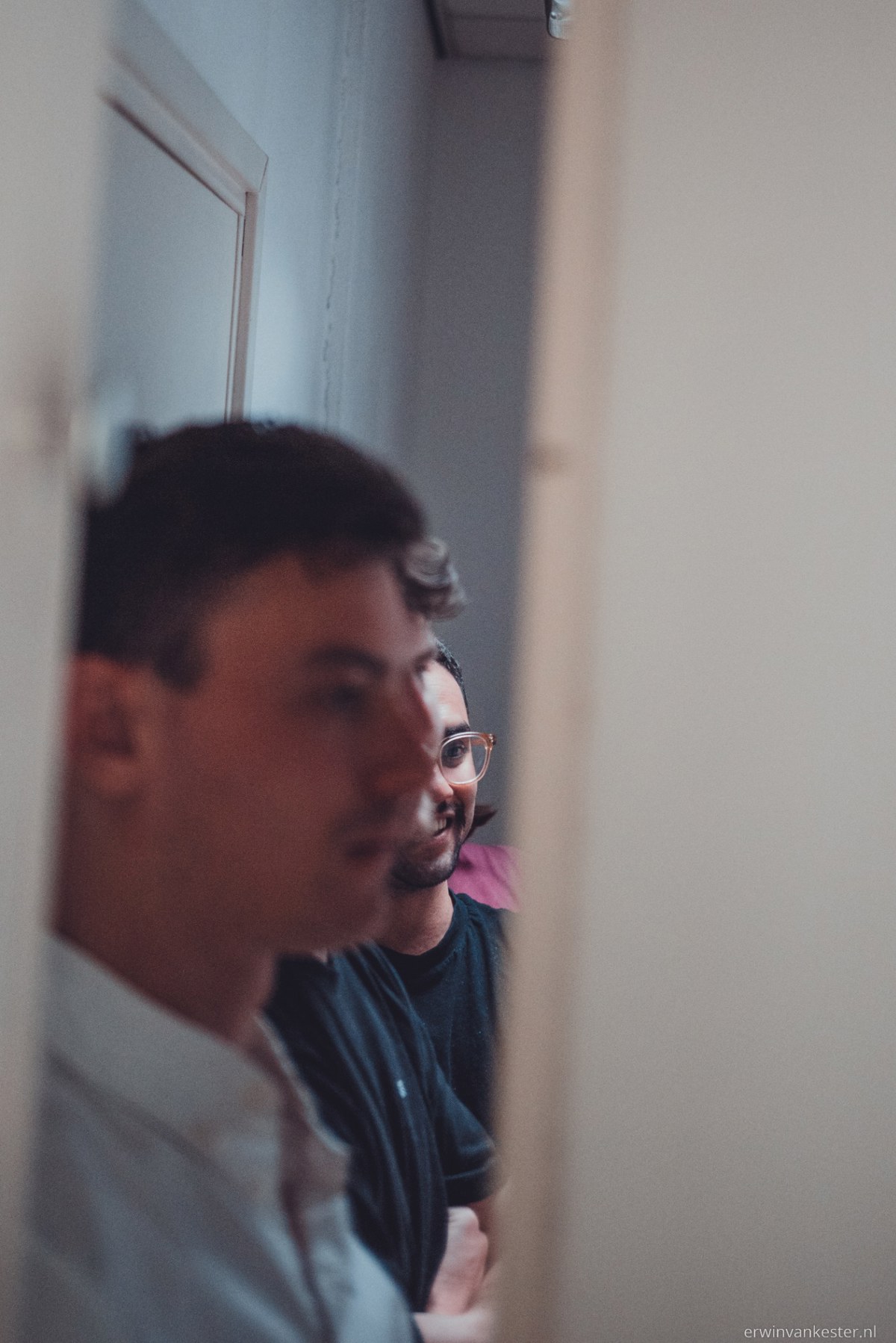 Twee mannen kijken geconcentreerd opzij vanuit een deuropening, waarbij de voorste onscherp is en de man met bril op de achtergrond scherp in beeld staat.