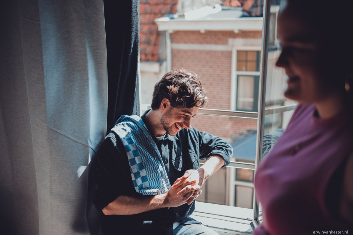 Lachende man met krullend haar zit bij een raam iets in zijn handen te bekijken, terwijl een vrouw in het paars op de voorgrond onscherp meekijkt.