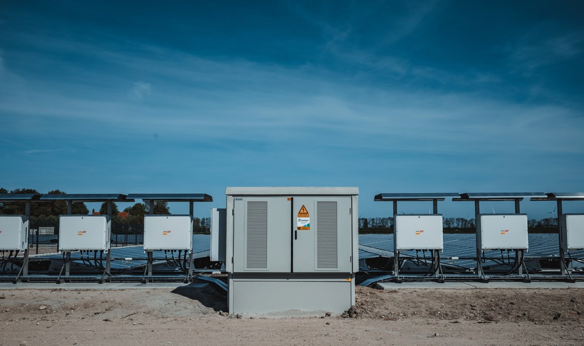 Een rij grijze technische kasten van een zonnepark staat opgesteld voor de zonnepanelen onder een strakblauwe lucht.