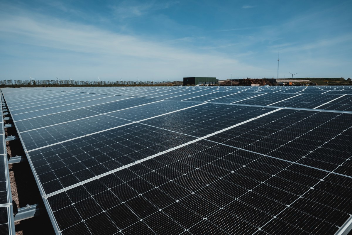 Rijen zwarte zonnepanelen strekken zich uit onder een strakblauwe lucht, met op de achtergrond een bedrijfsgebouw en een windturbine.