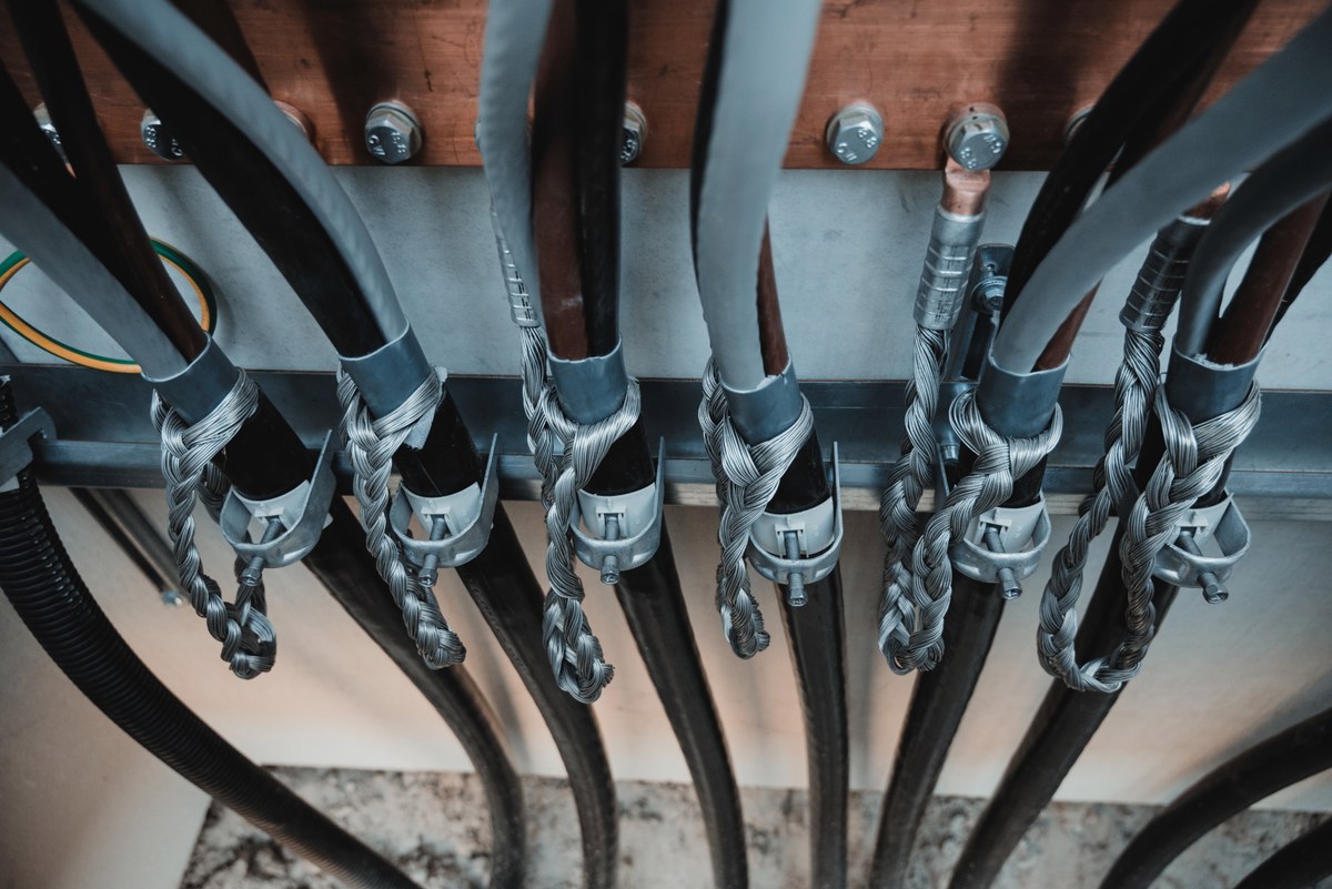 Rijen dikke zwarte kabels met gevlochten kabelkousen en metalen trekontlastingen, bevestigd aan een houten balk in een technische installatieruimte.