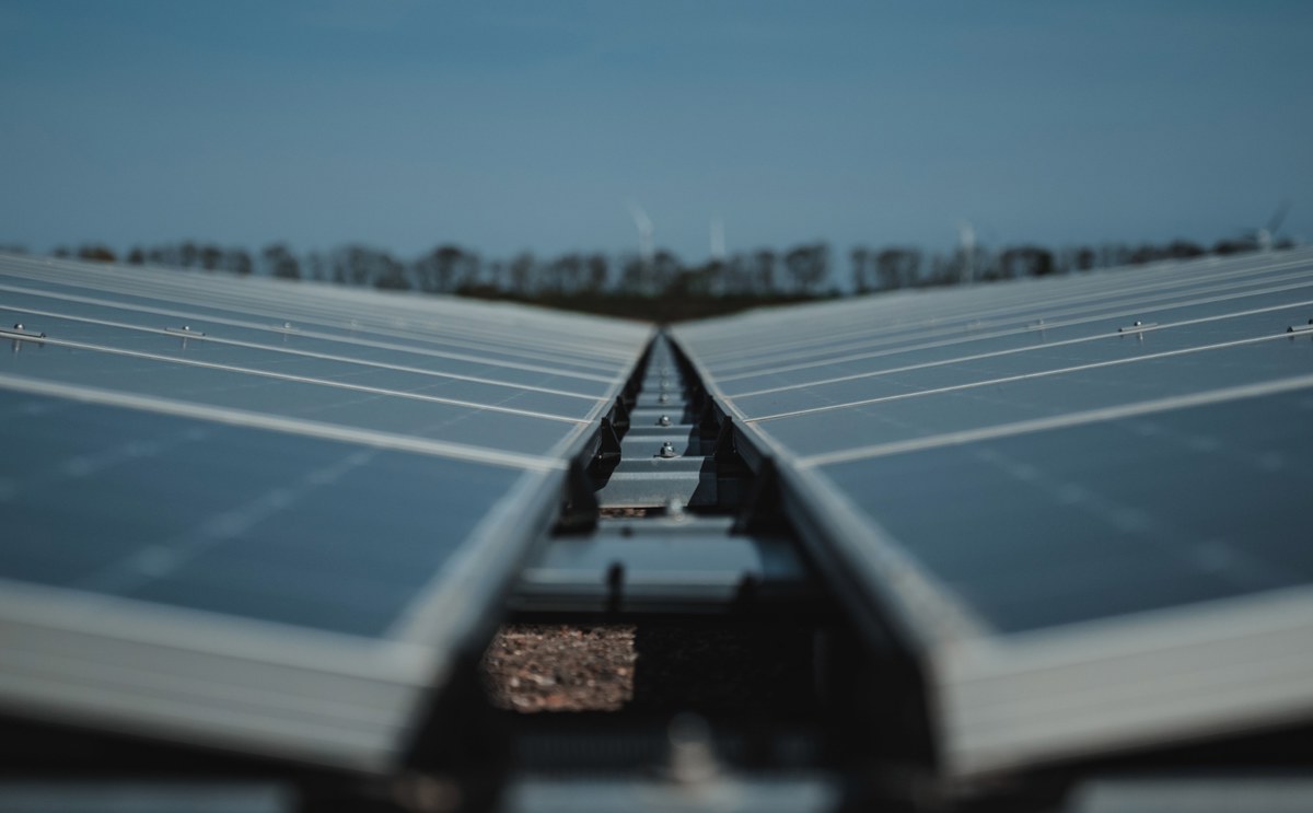 Twee rijen zonnepanelen lopen in een V-vorm naar elkaar toe onder een blauwe lucht, met in de verte windturbines en een bomenrij.
