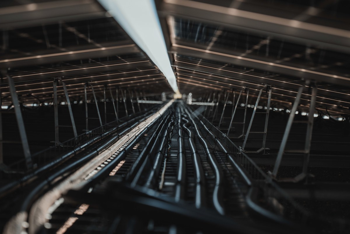 Onderaanzicht van een rij zonnepanelen op een frame van donkere metalen stangen en kabels, met een streep daglicht tussen de panelen.
