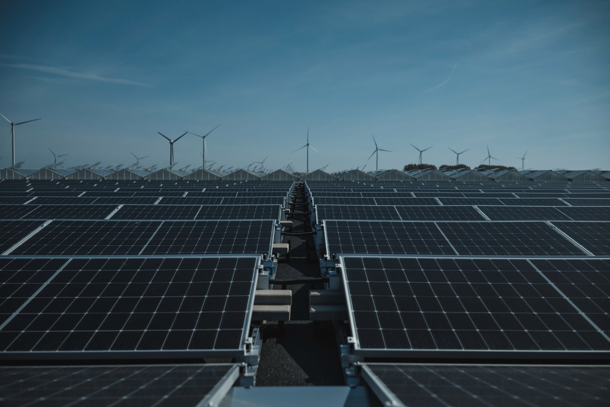 Rijen zonnepanelen strekken zich uit tot de horizon, waar windturbines onder een strakblauwe hemel oprijzen boen een groen heuvellandschap.