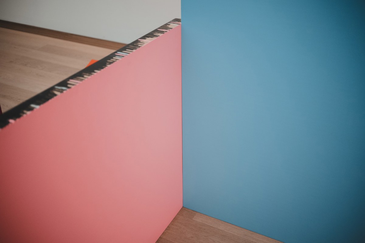 Een roze en een blauw kleurvlak vormen samen een hoek boven een houten vloer, met daarachter een witte muur en een streep gestructureerd materiaal langs de bovenrand.