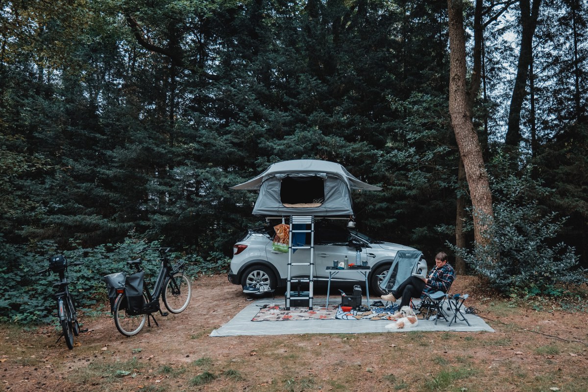Op een beboste kampeerplek staat een auto met opgezette daktent, ernaast een fiets en een persoon op een campingstoel op een picknickkleed.