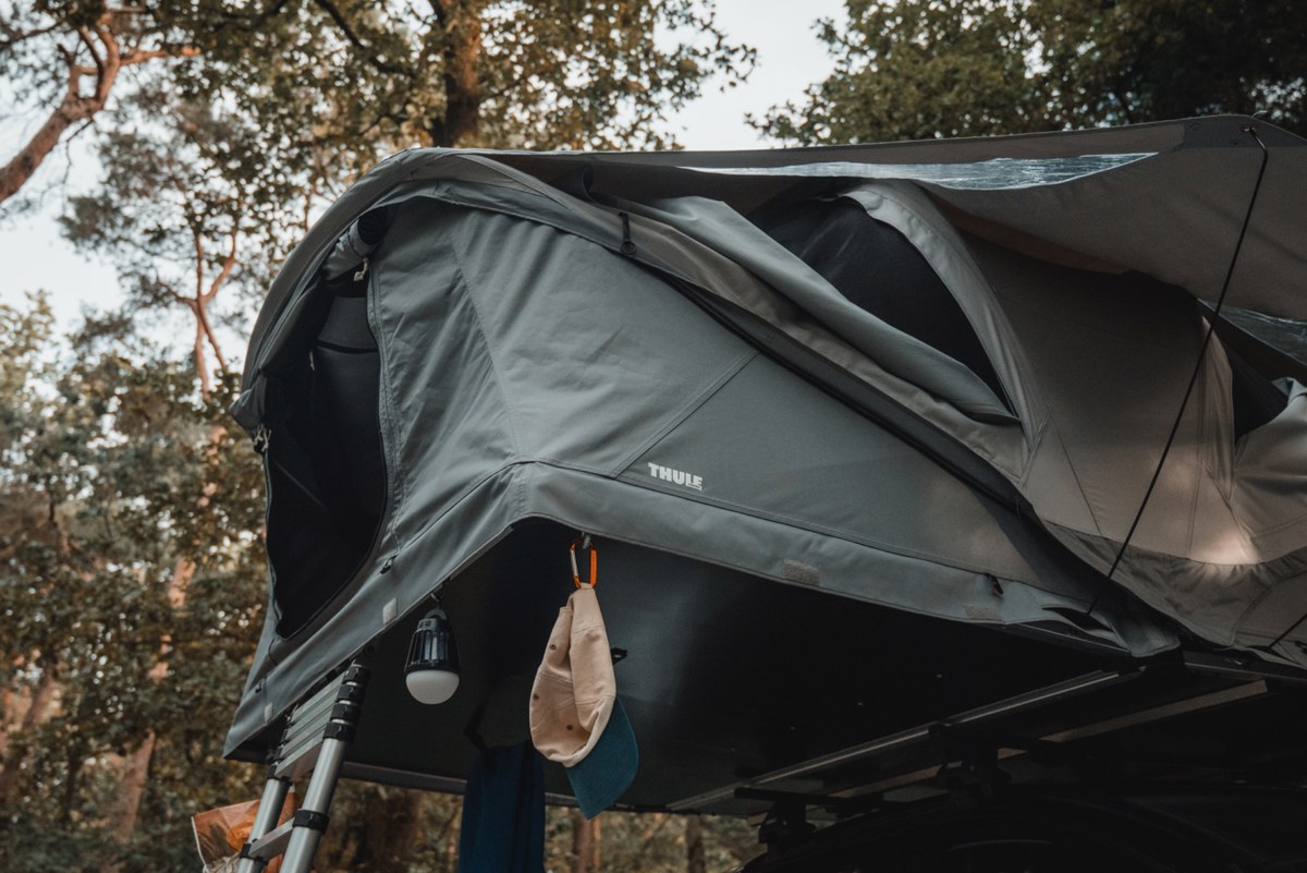 Een donkergrijze Thule Approach-daktent staat opgeklapt op een voertuig in het bos, met een hangende lamp en een petje aan haken en een ladder ernaast.