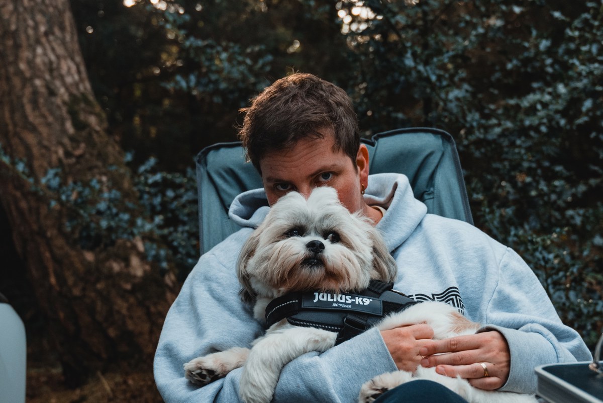 Een vrouw (Veerle) in een lichtblauwe hoodie zit in een campingstoel en houdt een hond (Kip) met Julius-K9-tuigje beschermend tegen zich aan tussen de bomen.