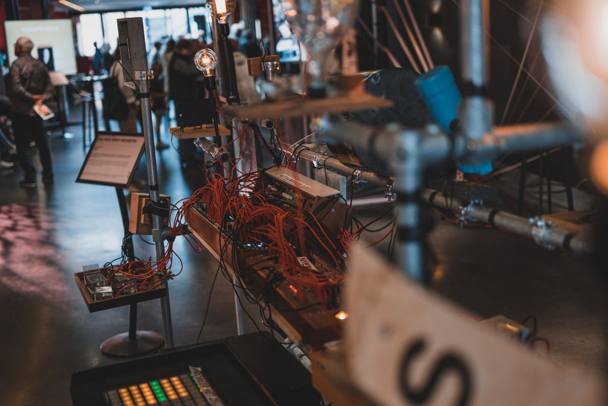 Interactieve robotinstallatie met een wirwar van oranje kabels, elektronica en gloeilampen in een museumzaal waar bezoekers rondlopen.