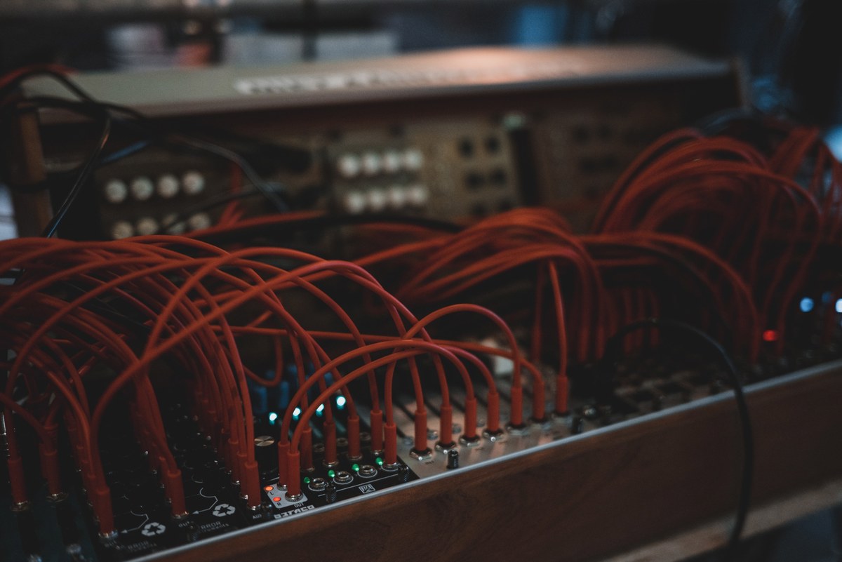 Talrijke rode patchkabels verbinden de aansluitingen van een modulaire synthesizer, waarvan enkele groene en rode lampjes oplichten in het donker.