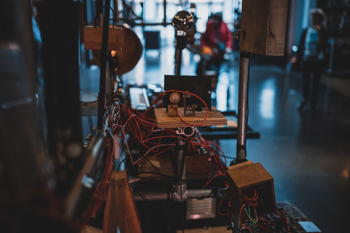 Zelfgebouwde elektronische installatie met een wirwar aan rode kabels en houten onderdelen op een tafel in een industrieel ogende ruimte met wazige figuren op de achtergrond.