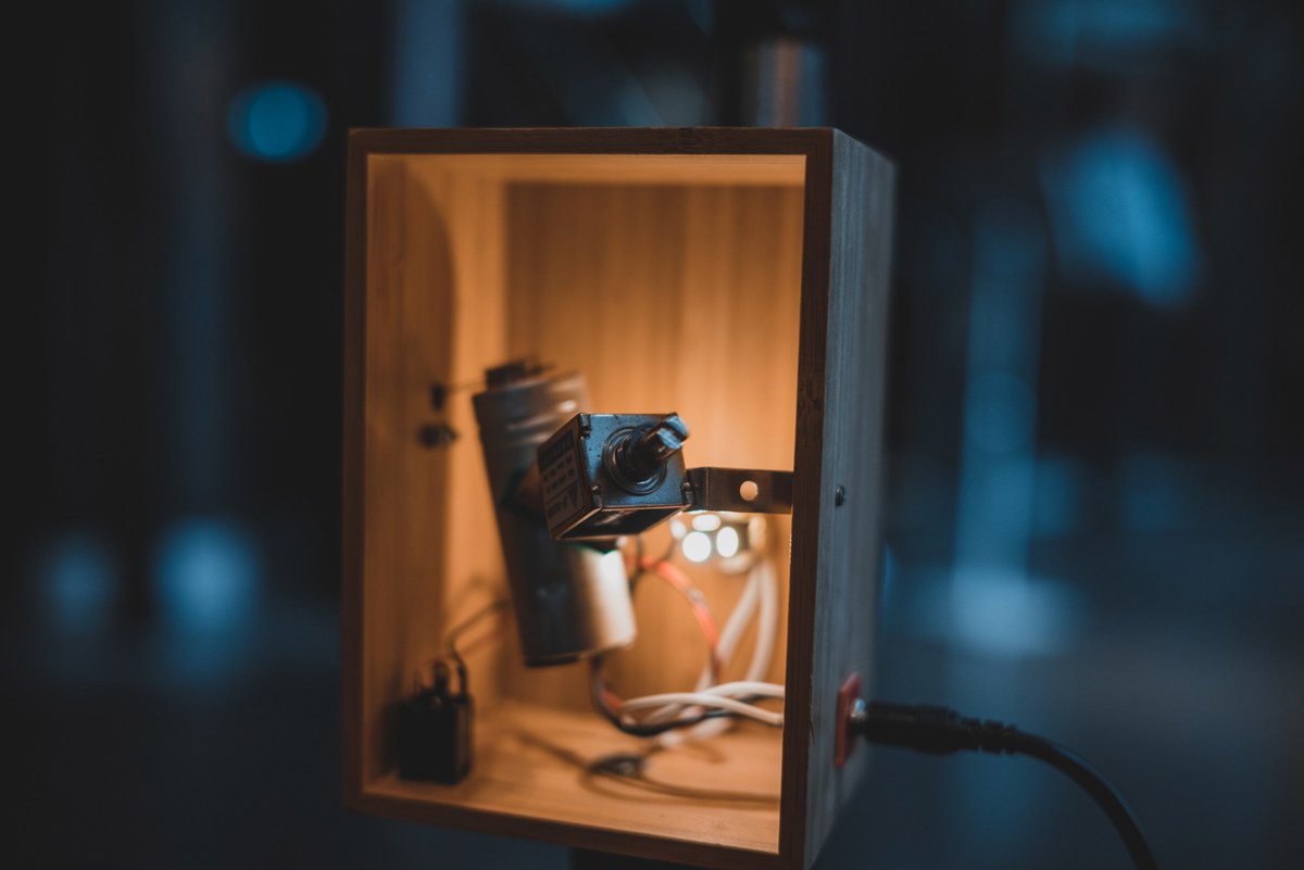 In een warm verlichte houten kist staat een klein cameramodule op een servomotor, met kabels die naar buiten lopen tegen een donkerblauwe achtergrond.