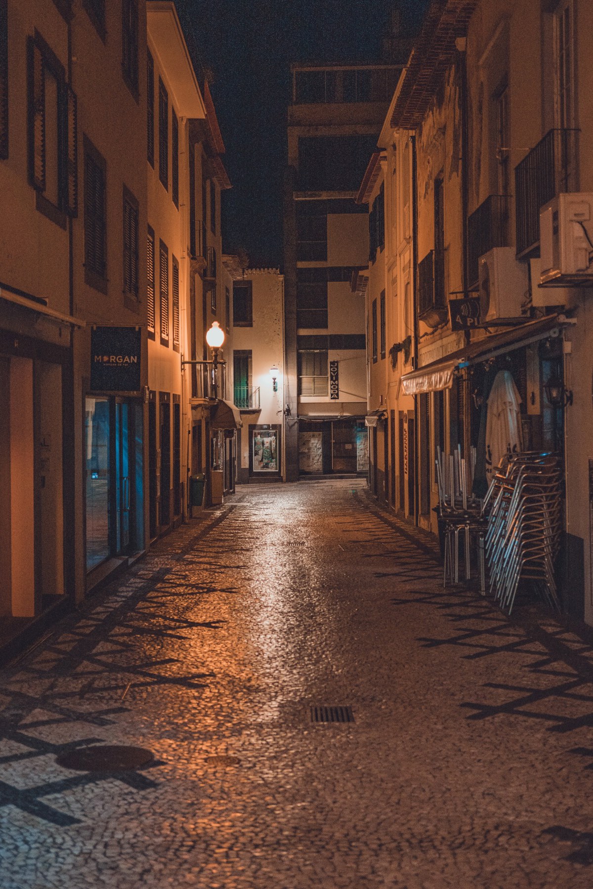 Een verlaten, geplaveide steeg in Funchal bij nacht, verlicht door een straatlantaarn, met opgestapelde terrasstoelen en warm gloeiende gevels.