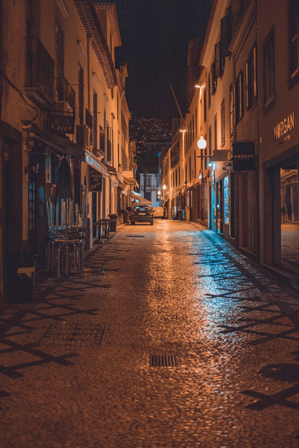 Een verlaten geplaveide straat in Funchal glinstert 's nachts onder oranje straatlantaarns, geflankeerd door winkels en een geparkeerde auto in de verte.