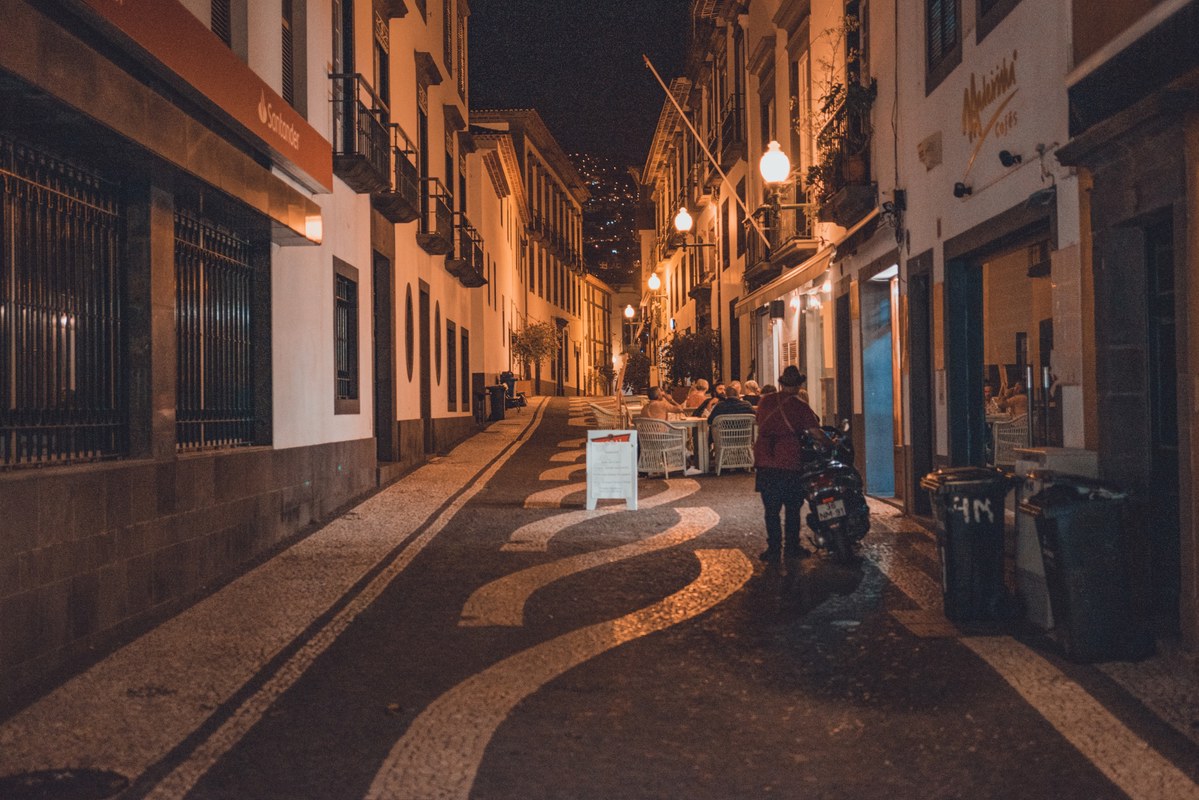 Een smalle geplaveide straat in Funchal bij nacht, verlicht door straatlantaarns, met terrasjes, een geparkeerde scooter en enkele voetgangers.