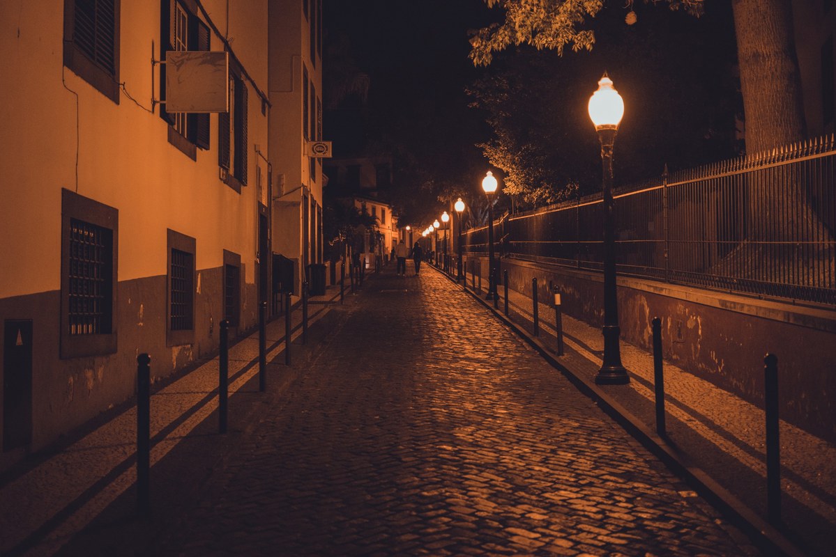Een verlaten geplaveide straat 's nachts, verlicht door warm gloeiende lantaarns langs een ijzeren hek en oude gebouwen aan weerszijden.