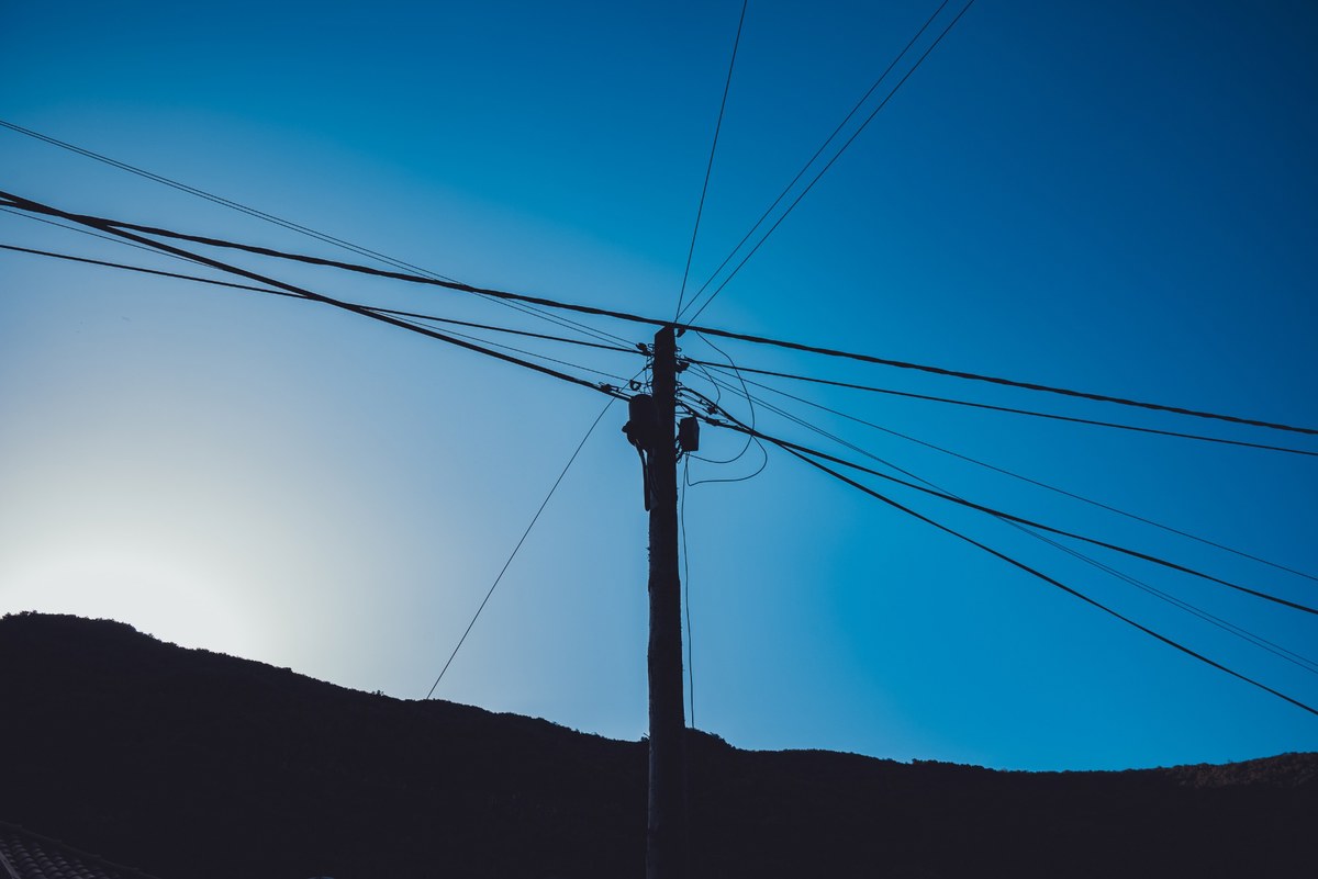 Een silhouet van een houten elektriciteitspaal met talrijke uitwaaierende draden tegen een diepblauwe avondlucht boven een donkere heuvel.
