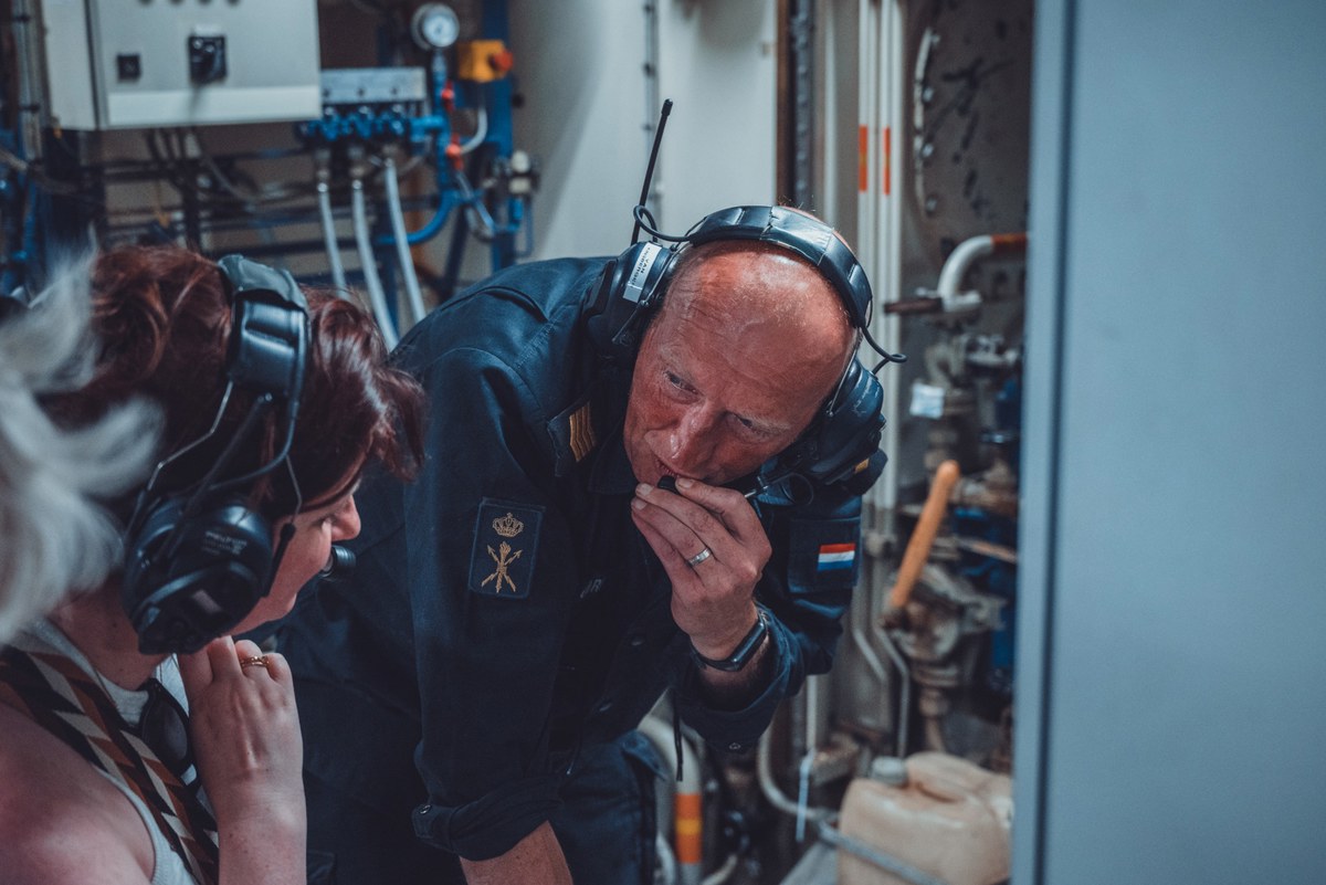 Marineofficier met gehoorbescherming buigt zich voorover en spreekt in een intercom in een machineruimte, terwijl een collega naast hem meeluistert.