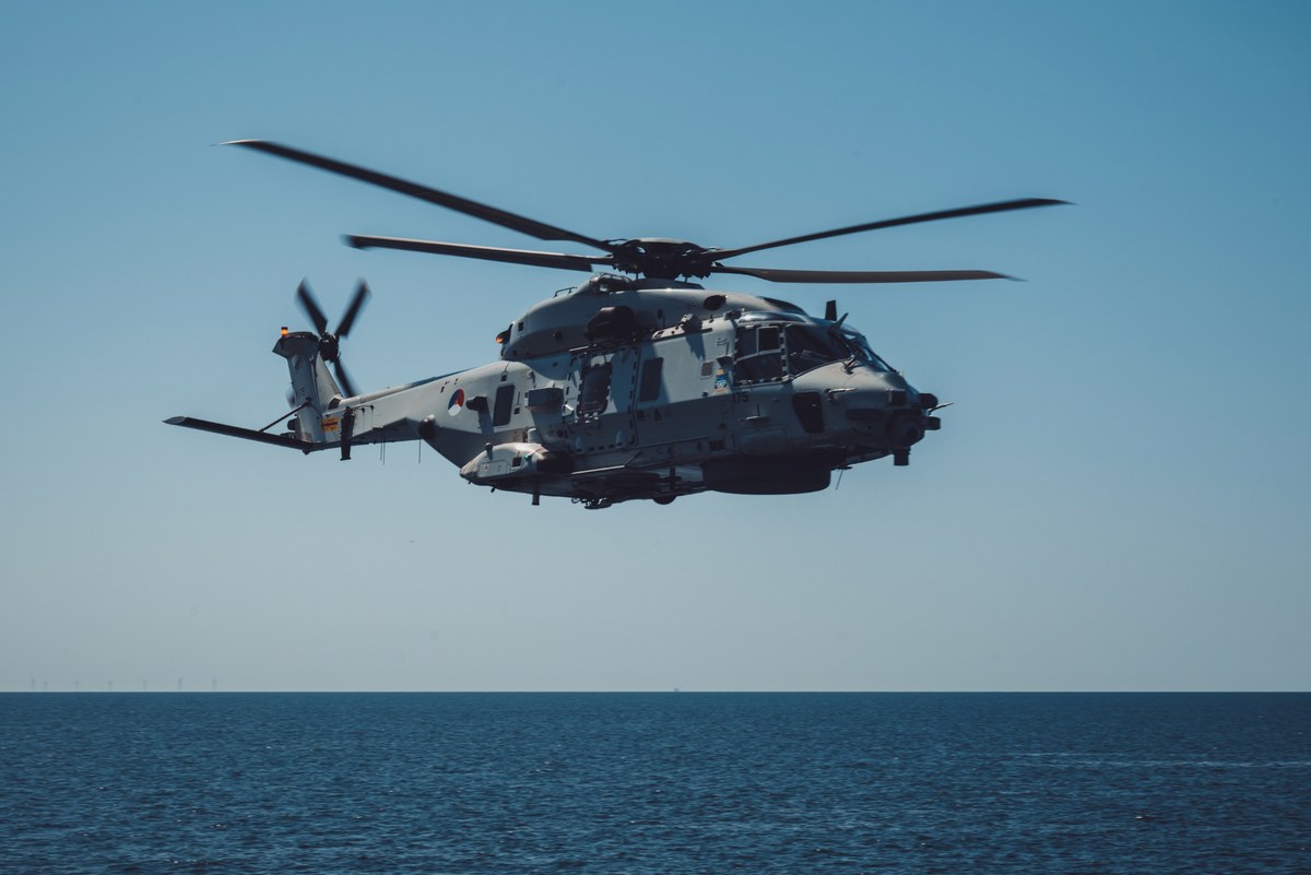 Een grijze militaire NH90-helikopter vliegt laag boven een kalme, diepblauwe zee onder een strakblauwe hemel, met windturbines vaag aan de horizon.