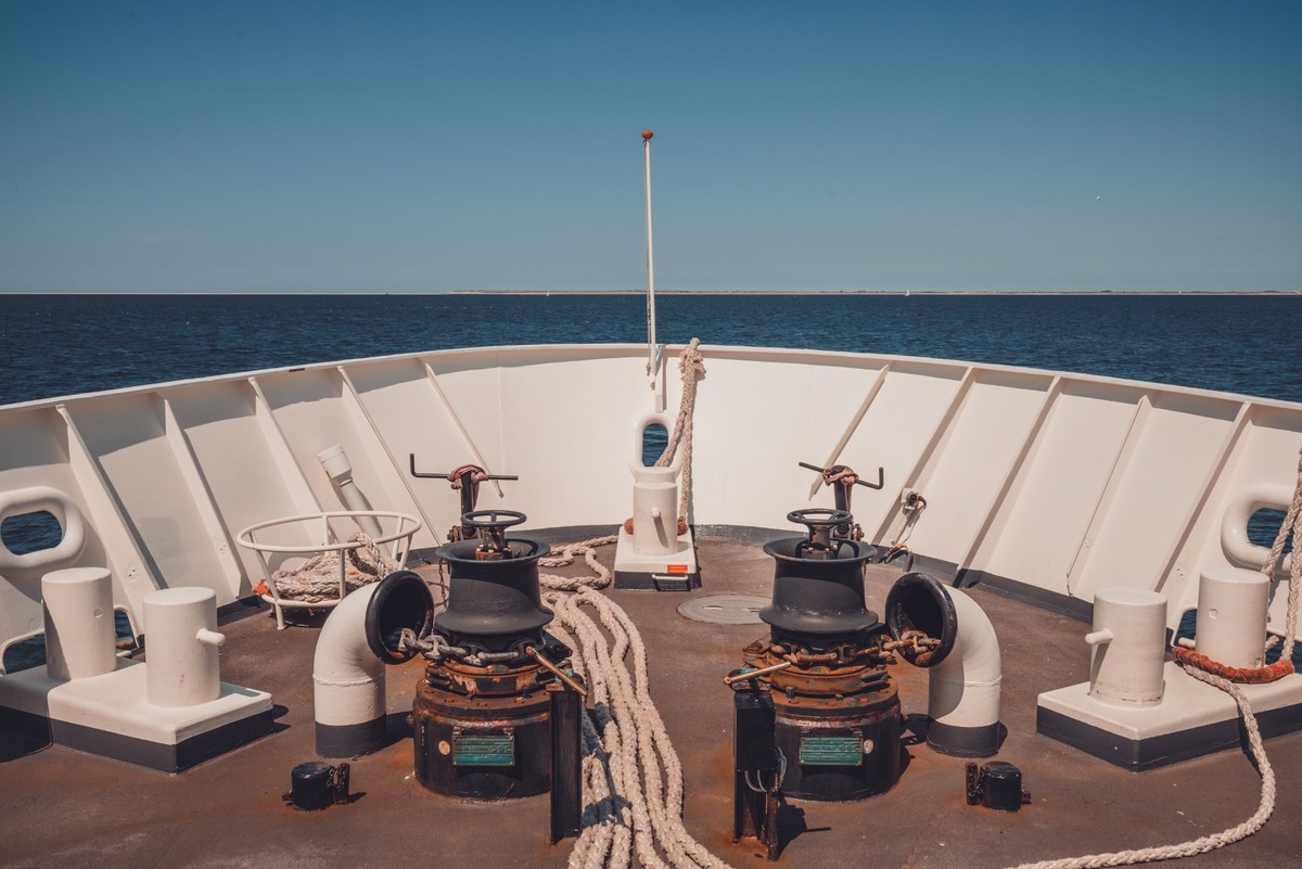 Het voordek van een schip met twee zwarte ankerlieren, opgerolde meertrossen en een witte reling, met daarachter een kalme blauwe zee onder een strakblauwe hemel.