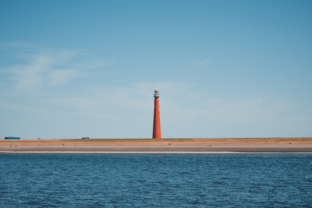 Een roodgekleurde vuurtoren staat op een smalle zandstrook tussen het water en de strakblauwe lucht, gezien vanaf de overkant van het water.