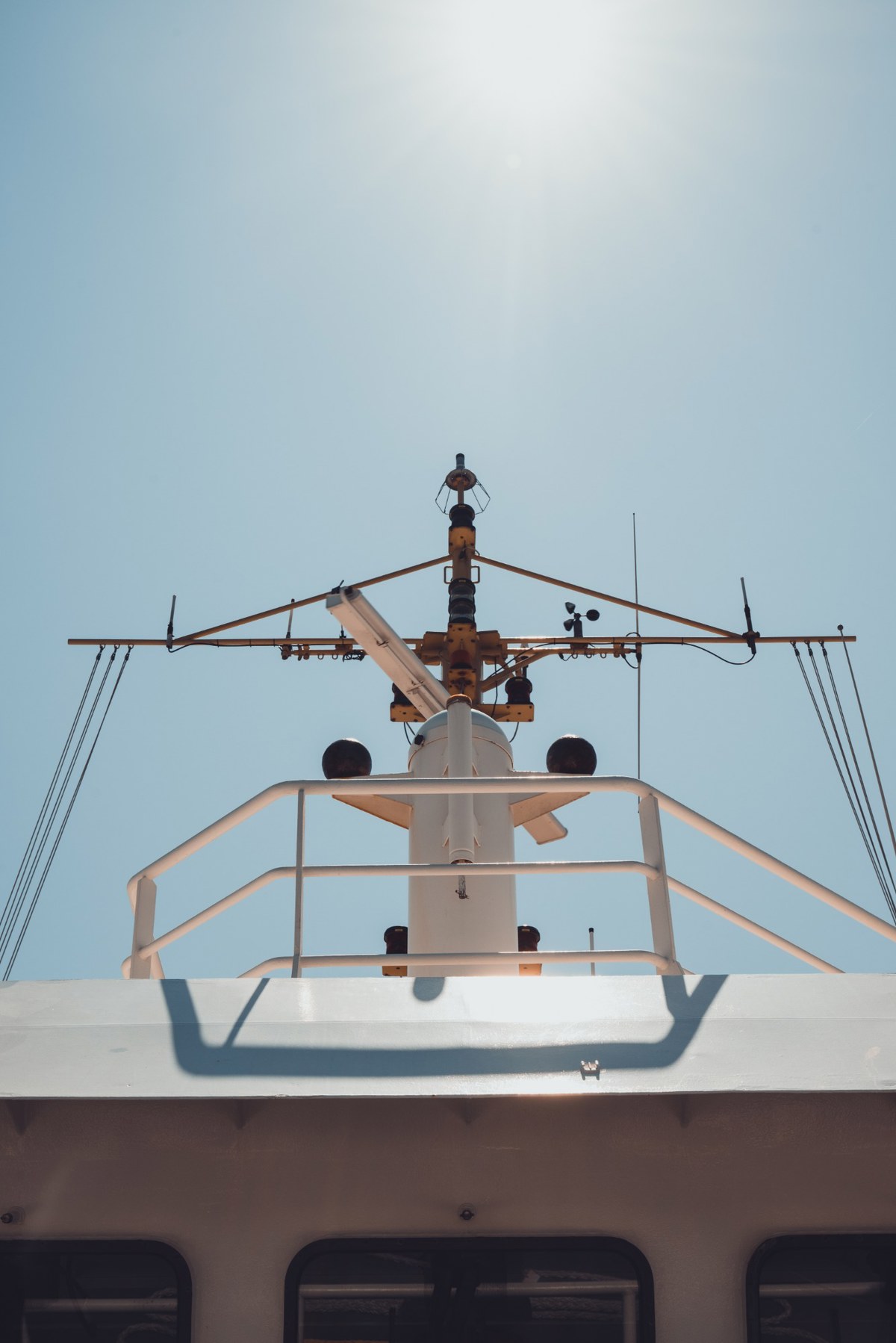 De mast met navigatieapparatuur en antennes van een schip steekt tegen een strakblauwe hemel omhoog, boven een witte reling en dekopbouw.