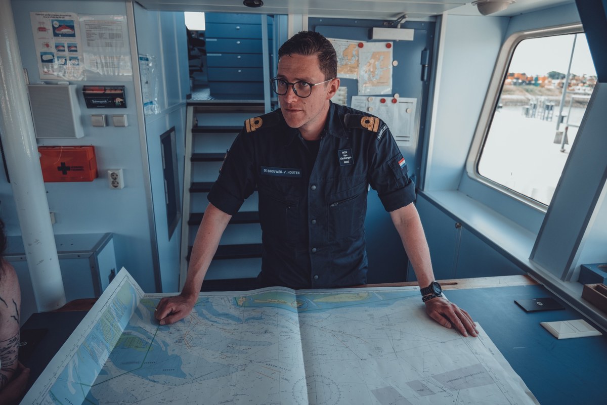 Een marineofficier met bril buigt zich over een zeekaart op de brug van een schip en kijkt geconcentreerd opzij tijdens de navigatie.