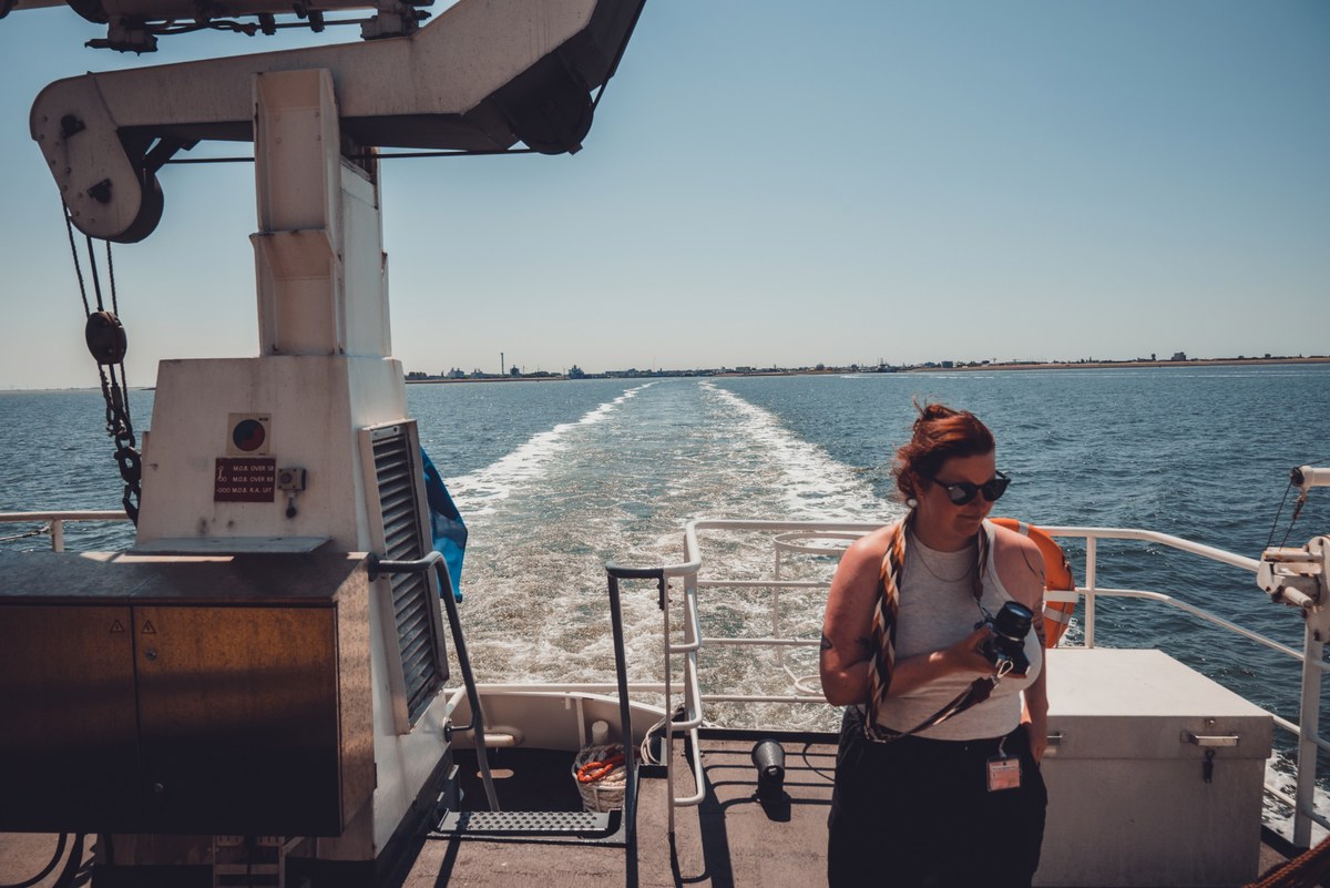 Een vrouw met zonnebril en camera staat op het achterdek van een schip, terwijl het kielzog een lang spoor door het kalme water trekt.