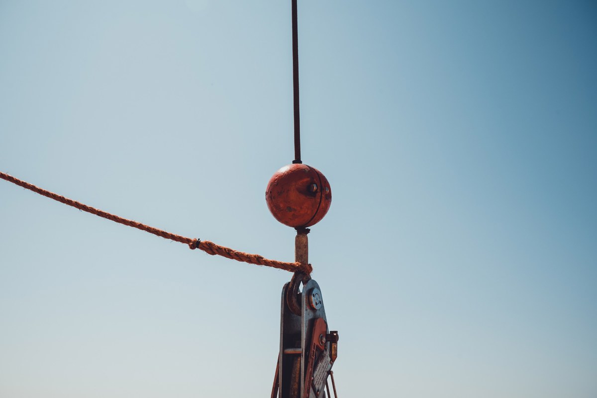 Een verweerde roestbruine haakblok met kraanhaak hangt aan een kabel tegen een strakblauwe lucht, met een gerafeld touw dat schuin het beeld in loopt.