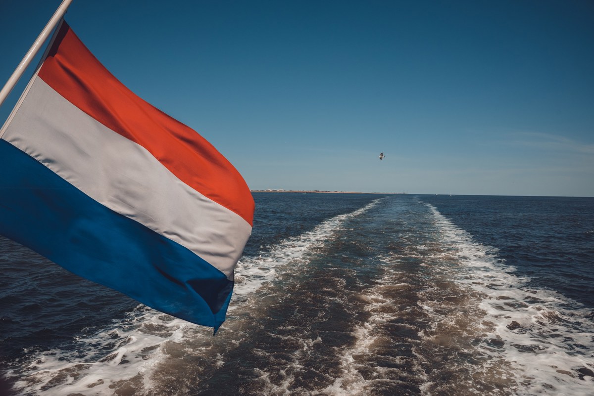 De Nederlandse driekleur wappert aan de achterzijde van een varend schip terwijl het schuimende kielzog een spoor trekt over de blauwe zee.