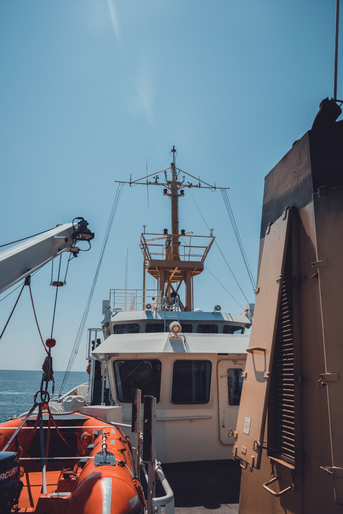 Vanaf het dek van een schip zie je de witte brug met mast en antennes, een oranje reddingssloep en een crane onder een strakblauwe hemel boven zee.