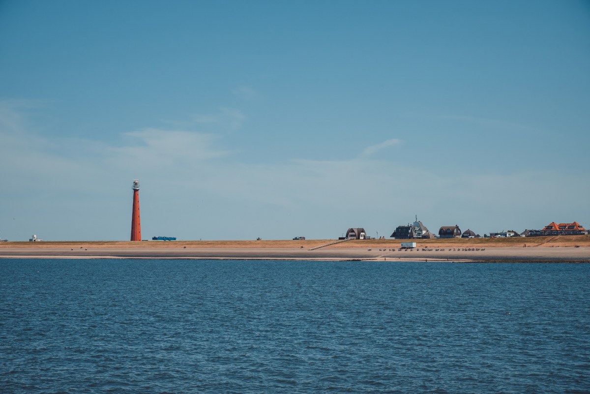 Een rode vuurtoren rijst op boven een langgerekte kustlijn met verspreide huizen, gezien vanaf het kalme water onder een strakblauwe lucht.