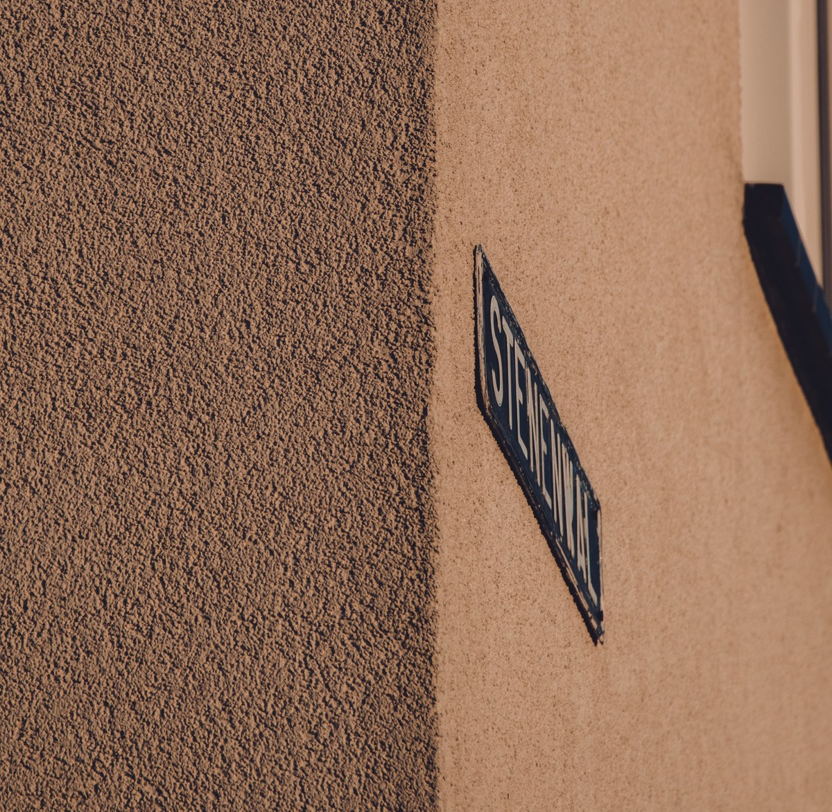 Een blauw geëmailleerd straatnaambordje op de hoek van een gebouw met ruwe pleisterwand en een gladde, warmbeige muur.