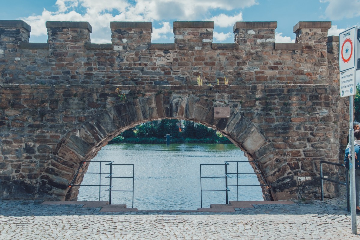 Een oude gekanteelde stenen muur met een grote boog waardoorheen water zichtbaar is, met op de voorgrond kinderkopjes en een metalen hekwerk.