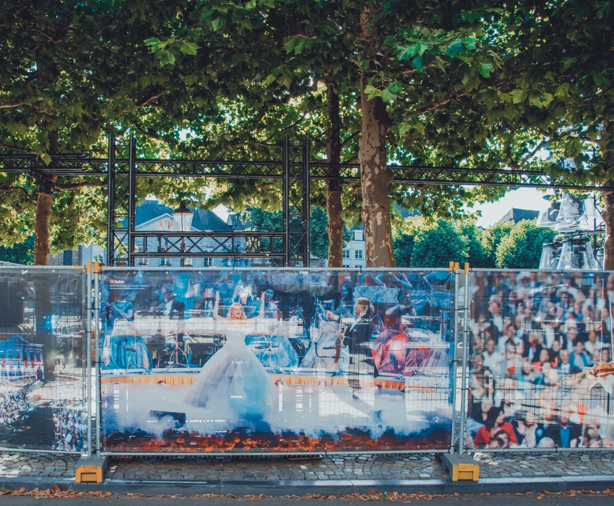 Grote fotobanners van een operavoorstelling met een vrouw in witte jurk hangen aan bouwhekken onder rijen bomen op een Maastrichts plein.