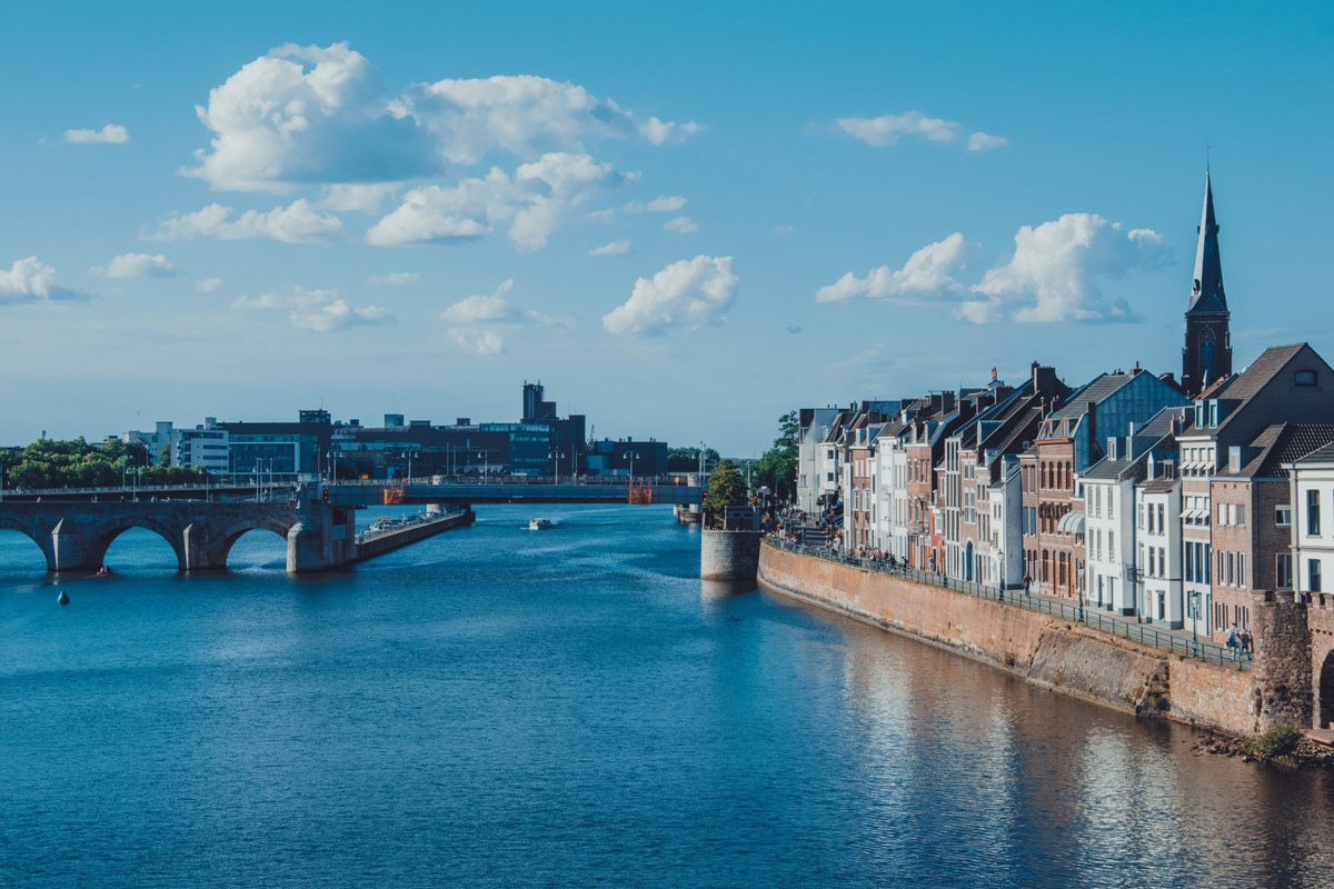 Rijtjeshuizen langs de Maas met een oude boogbrug op de achtergrond en een kerktoren die boven de skyline uitsteekt onder een blauwe lucht.