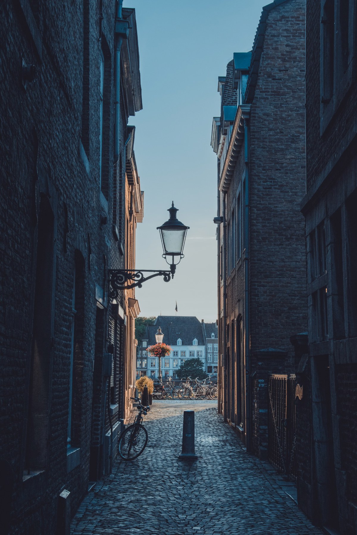 Een smalle geplaveide steeg tussen bakstenen huizen met een sierlijke lantaarn en fiets, uitkijkend op een zonovergoten plein met historische gevels.