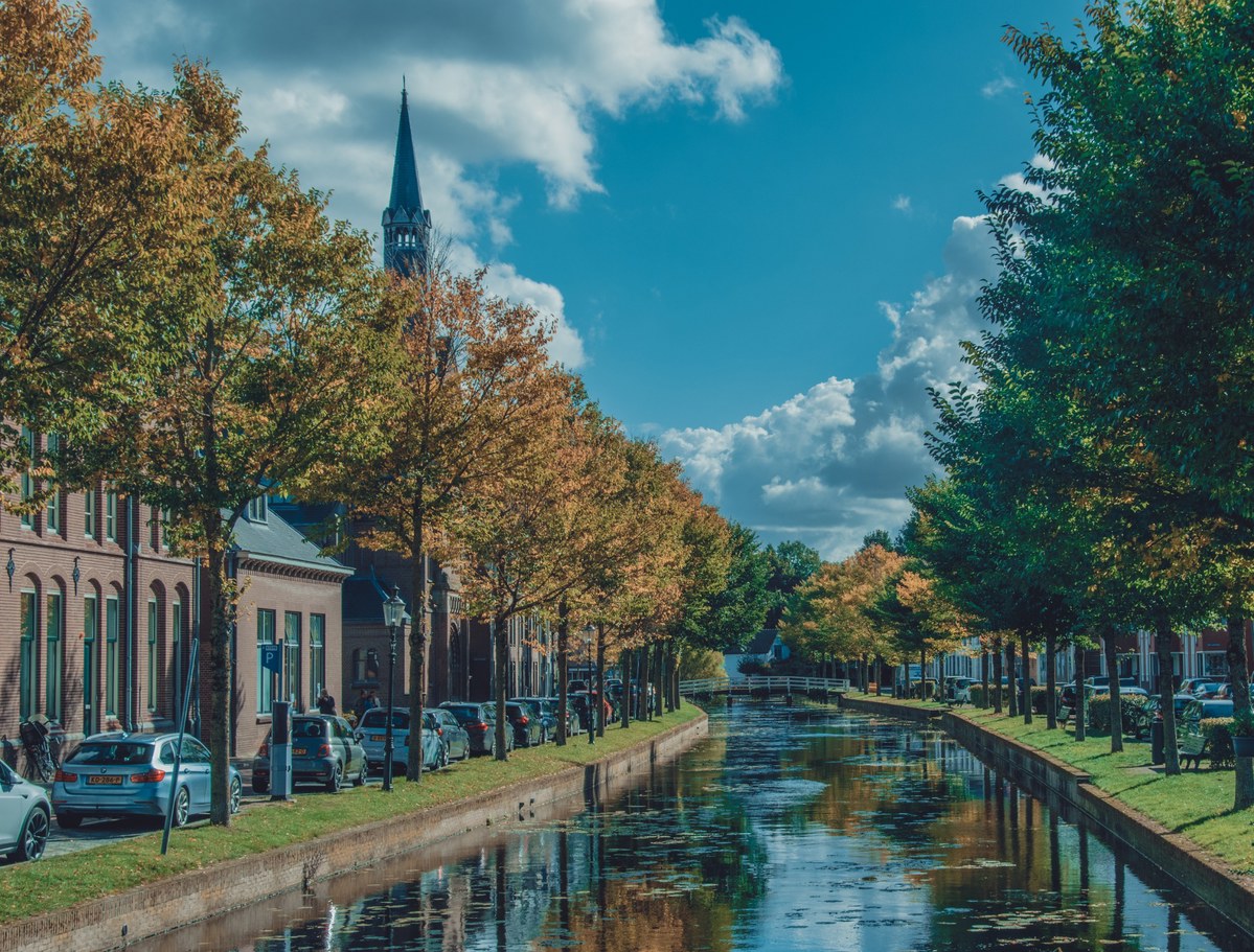 Een smal grachtje doorsnijdt een historisch straatje met bomen en geparkeerde auto's, terwijl een kerktoren oprijst tegen een blauwe wolkenlucht.