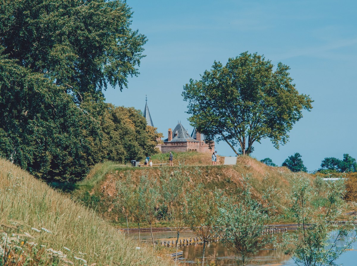 Middeleeuws kasteel met torens en spitsen steekt boven groene bomen en een begroeide wal uit, met wandelaars op het pad en water op de voorgrond.
